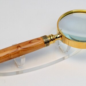 May include: A magnifying glass with a gold-colored rim and handle accents. The handle is made of light-colored wood with a dark tip. The magnifying glass is resting on a clear stand.
