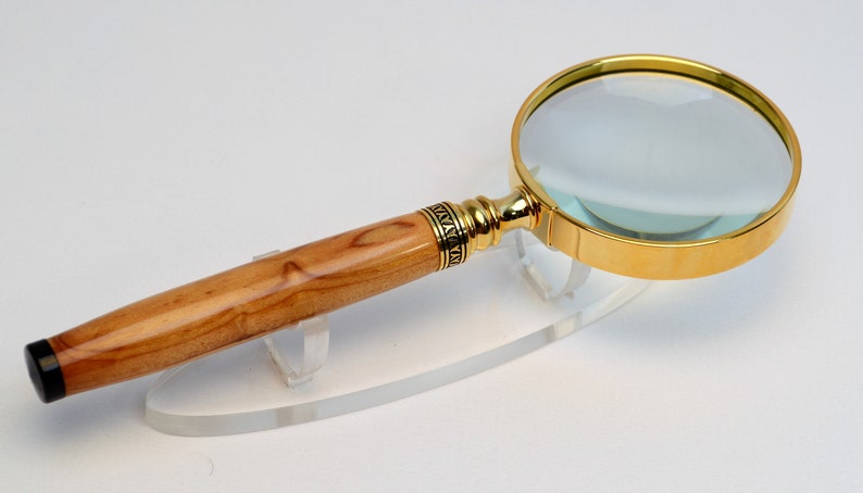 May include: A magnifying glass with a gold-colored rim and handle accents. The handle is made of light-colored wood with a dark tip. The magnifying glass is resting on a clear stand.