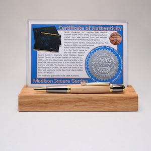 May include: A wooden pen with a light-colored barrel, silver accents, and gold trim, displayed on a small wooden block. A certificate of authenticity is behind the pen, with text that reads "Madison Square Garden".