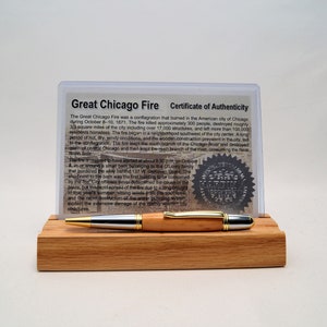 May include: A wooden pen with silver and gold accents rests on a wooden stand, in front of a certificate of authenticity. The certificate details the Great Chicago Fire of 1871. The pen is approximately 5.5 inches long.