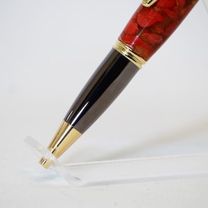 May include: A close-up of a handcrafted pen. The pen features a red stone resin barrel with gold accents and a dark gray metal body. The pen is displayed on a clear acrylic stand, showcasing its design and craftsmanship.