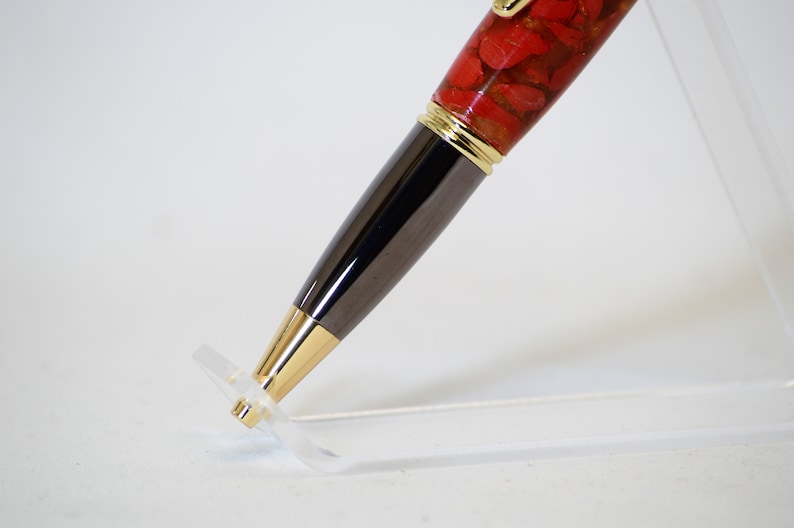 May include: A close-up of a handcrafted pen. The pen features a red stone resin barrel with gold accents and a dark gray metal body. The pen is displayed on a clear acrylic stand, showcasing its design and craftsmanship.