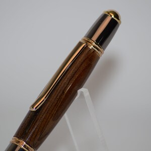 Puede incluir: Primer plano de un bol&iacute;grafo de lujo con cuerpo de madera y detalles dorados. El bol&iacute;grafo presenta un patr&oacute;n de vetas de madera marr&oacute;n oscuro, complementado con adornos dorados y una punta dorada. El bol&iacute;grafo se exhibe sobre un soporte acr&iacute;lico transparente.