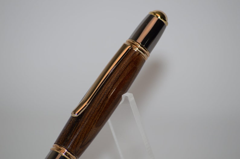 Puede incluir: Primer plano de un bol&iacute;grafo de lujo con cuerpo de madera y detalles dorados. El bol&iacute;grafo presenta un patr&oacute;n de vetas de madera marr&oacute;n oscuro, complementado con adornos dorados y una punta dorada. El bol&iacute;grafo se exhibe sobre un soporte acr&iacute;lico transparente.