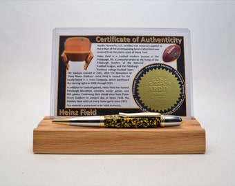 Könnte beinhalten: Ein Stift mit einem schwarz-gelb gemusterten Schaft, silbernen Akzenten und einer goldfarbenen Spitze liegt auf einem Holzständer. Ein Echtheitszertifikat wird hinter dem Stift mit dem Text "Heinz Field" angezeigt.