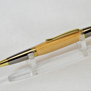 Peut inclure: Un stylo en bois avec des d&eacute;tails dor&eacute;s et argent&eacute;s. Le stylo a un corps en bois clair, un clip dor&eacute; et une pointe et une extr&eacute;mit&eacute; argent&eacute;es. Il est pr&eacute;sent&eacute; sur un support en plastique transparent sur fond blanc.