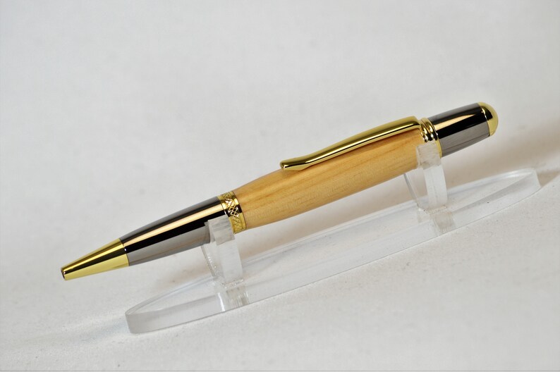 Peut inclure: Un stylo en bois avec des d&eacute;tails dor&eacute;s et argent&eacute;s. Le stylo a un corps en bois clair, un clip dor&eacute; et une pointe et une extr&eacute;mit&eacute; argent&eacute;es. Il est pr&eacute;sent&eacute; sur un support en plastique transparent sur fond blanc.