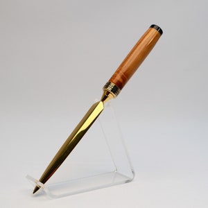 May include: A handcrafted letter opener with a polished gold blade and a wooden handle. The handle features a light brown wood grain with a decorative gold band near the blade. The letter opener is displayed on a clear acrylic stand.