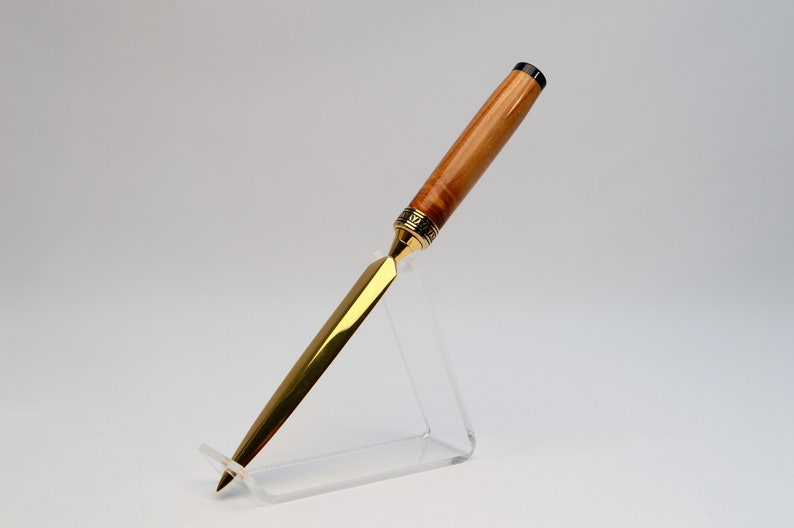 May include: A handcrafted letter opener with a polished gold blade and a wooden handle. The handle features a light brown wood grain with a decorative gold band near the blade. The letter opener is displayed on a clear acrylic stand.