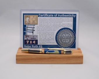 Puede incluir: Un bolígrafo con detalles en tono dorado y un cuerpo de madera con un gráfico de Babe Ruth, exhibido sobre un soporte de madera rectangular frente a una tarjeta de certificado de autenticidad que detalla el uso de reliquias de camiseta y bate.