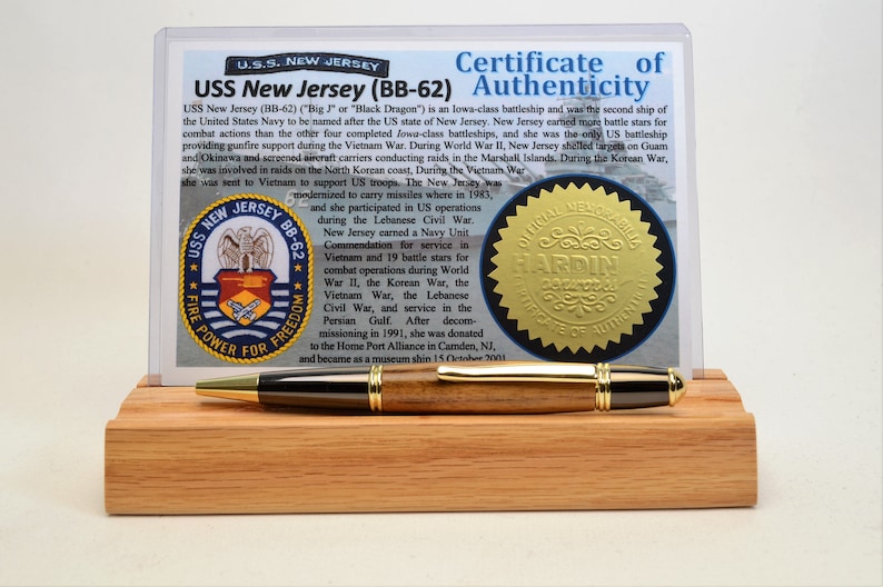 May include: A wooden pen with gold accents rests on a wooden display stand, in front of a certificate of authenticity for the U.S.S. New Jersey (BB-62). The certificate includes text and a gold seal.