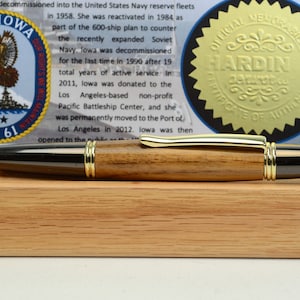 Puede incluir: Un bol&iacute;grafo de madera con detalles dorados y punta negra, exhibido sobre un soporte de madera. El bol&iacute;grafo est&aacute; junto a un documento con el texto "USS IOWA BB 61" y un sello dorado que dice "OFFICIAL MEMORABILIA HARDIN CERTIFICATE OF AUTHENTICITY."