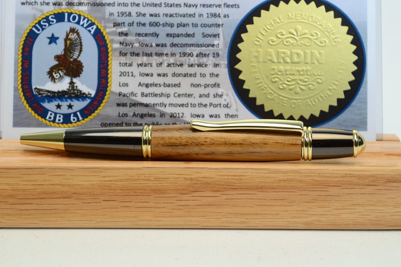 Puede incluir: Un bol&iacute;grafo de madera con detalles dorados y punta negra, exhibido sobre un soporte de madera. El bol&iacute;grafo est&aacute; junto a un documento con el texto "USS IOWA BB 61" y un sello dorado que dice "OFFICIAL MEMORABILIA HARDIN CERTIFICATE OF AUTHENTICITY."