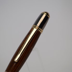 May include: A close-up of a wooden pen with gold-colored accents. The pen has a dark brown barrel and a black top with a gold-colored tip. The pen clip and trim are also gold-colored. The pen is set against a white background.