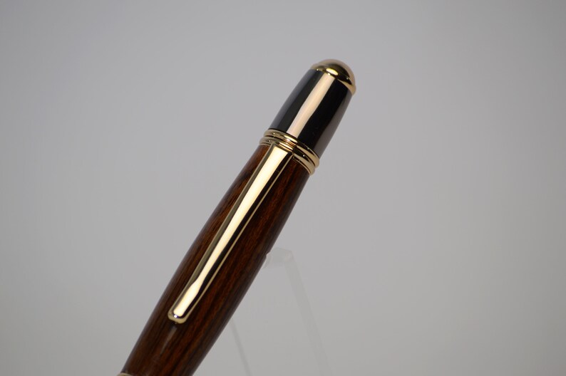 May include: A close-up of a wooden pen with gold-colored accents. The pen has a dark brown barrel and a black top with a gold-colored tip. The pen clip and trim are also gold-colored. The pen is set against a white background.