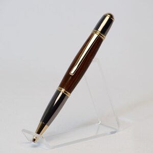 May include: A handcrafted pen with a dark wood barrel, accented with gold-colored trim and a black tip. The pen is displayed on a clear acrylic stand, showcasing its elegant design. The pen's length is approximately 6 inches.