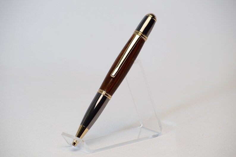 May include: A handcrafted pen with a dark wood barrel, accented with gold-colored trim and a black tip. The pen is displayed on a clear acrylic stand, showcasing its elegant design. The pen's length is approximately 6 inches.