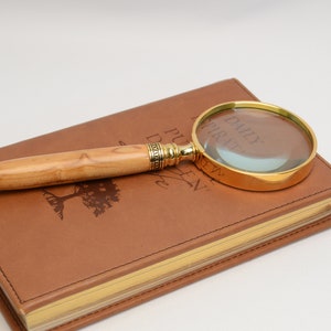 May include: A magnifying glass with a gold-colored frame and a light-colored wooden handle rests on a brown leather-bound journal. The journal has gold-colored edges and a red ribbon bookmark. The journal has the words "DAILY INSPIRATION" printed on the cover.