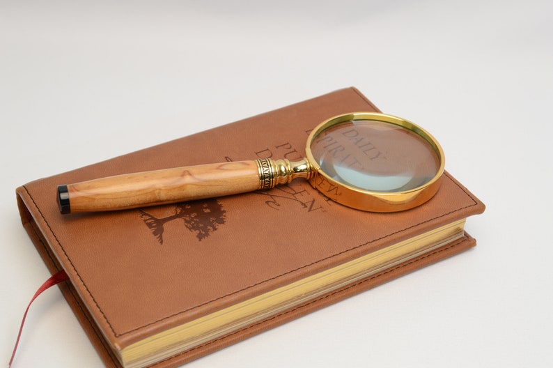 May include: A magnifying glass with a gold-colored frame and a light-colored wooden handle rests on a brown leather-bound journal. The journal has gold-colored edges and a red ribbon bookmark. The journal has the words "DAILY INSPIRATION" printed on the cover.