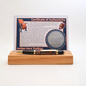 May include: A pen with a black barrel and orange and blue accents, resting on a wooden stand. Behind it is a certificate with text and a football illustration. The certificate is in a clear plastic holder.