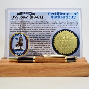 Puede incluir: Un bol&iacute;grafo de madera con detalles dorados descansa sobre un soporte de madera. Detr&aacute;s, un certificado de autenticidad para el USS Iowa (BB-61), un acorazado retirado. El certificado incluye el emblema del barco y detalles hist&oacute;ricos.