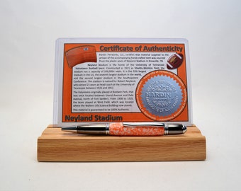 May include: A handcrafted pen with an orange and white speckled barrel, silver accents, and a black tip, displayed on a wooden stand. A certificate of authenticity for Neyland Stadium is behind the pen.