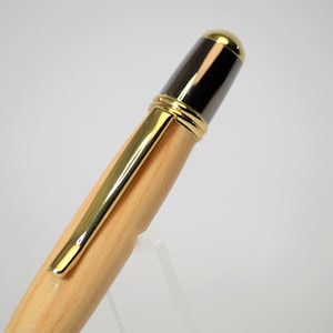 Peut inclure: Gros plan d'un stylo en bois avec des d&eacute;tails dor&eacute;s. Le stylo est dot&eacute; d'un clip dor&eacute; et d'une pointe dor&eacute;e. Le haut du stylo est noir et dor&eacute;. Le stylo est pr&eacute;sent&eacute; sur un support transparent.