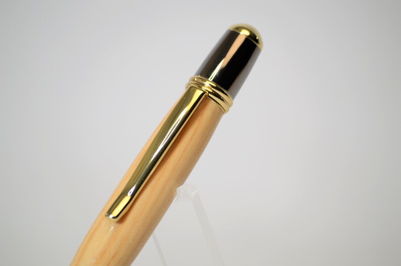 Peut inclure: Gros plan d'un stylo en bois avec des d&eacute;tails dor&eacute;s. Le stylo est dot&eacute; d'un clip dor&eacute; et d'une pointe dor&eacute;e. Le haut du stylo est noir et dor&eacute;. Le stylo est pr&eacute;sent&eacute; sur un support transparent.