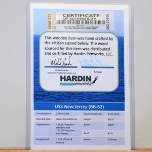 May include: A framed certificate of authenticity for a wooden item, featuring the USS New Jersey (BB-62). The certificate includes details like commissioning and decommissioning dates, class, length (887 ft), and displacement.