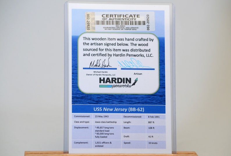 May include: A framed certificate of authenticity for a wooden item, featuring the USS New Jersey (BB-62). The certificate includes details like commissioning and decommissioning dates, class, length (887 ft), and displacement.