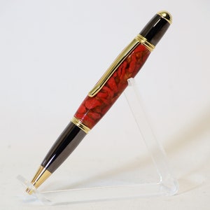 May include: A handcrafted pen with a red and gold patterned barrel, black accents, and gold-colored trim. The pen is displayed on a clear acrylic stand, showcasing its elegant design. A stylish writing instrument.