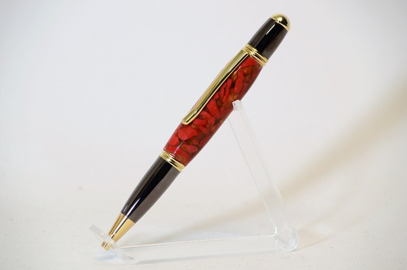 May include: A handcrafted pen with a red and gold patterned barrel, black accents, and gold-colored trim. The pen is displayed on a clear acrylic stand, showcasing its elegant design. A stylish writing instrument.