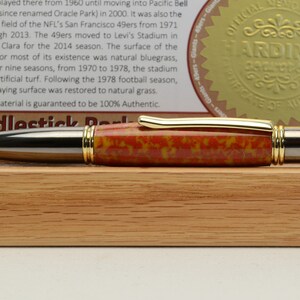 May include: A handcrafted pen with a red, yellow, and black patterned barrel, gold accents, and a silver tip. The pen is displayed on a wooden block, with a certificate and text in the background.