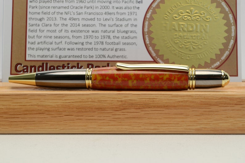 May include: A handcrafted pen with a red, yellow, and black patterned barrel, gold accents, and a silver tip. The pen is displayed on a wooden block, with a certificate and text in the background.