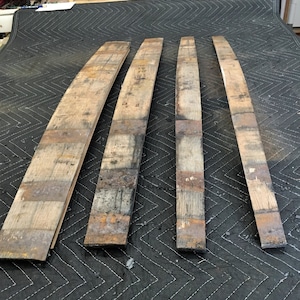 K&ouml;nnte beinhalten: Vier wiederverwertete Holzdauben, jede mit einer gebogenen Form. Die Dauben bestehen aus verwittertem Holz mit sichtbarer Maserung und Farbvariationen, darunter Braunt&ouml;ne und Grau. Sie sind auf einer dunklen, strukturierten Oberfl&auml;che angeordnet.