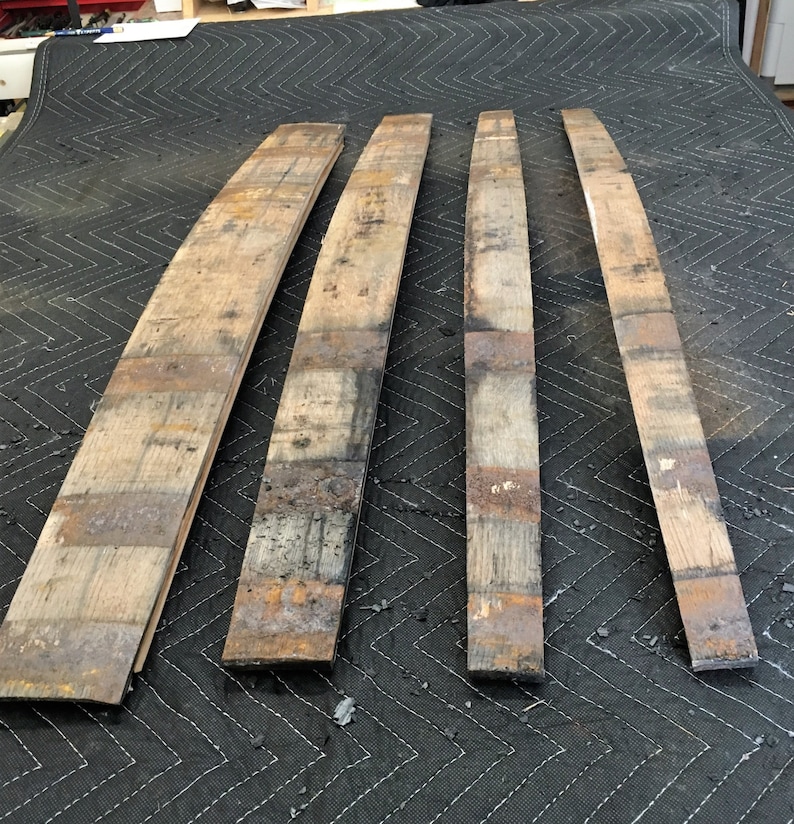 K&ouml;nnte beinhalten: Vier wiederverwertete Holzdauben, jede mit einer gebogenen Form. Die Dauben bestehen aus verwittertem Holz mit sichtbarer Maserung und Farbvariationen, darunter Braunt&ouml;ne und Grau. Sie sind auf einer dunklen, strukturierten Oberfl&auml;che angeordnet.
