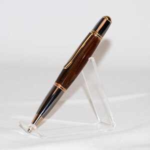 Puede incluir: Pluma de madera artesanal con cuerpo marr&oacute;n oscuro y detalles dorados. La pluma presenta un dise&ntilde;o elegante con punta negra y clip dorado. Se exhibe sobre un soporte de acr&iacute;lico transparente, mostrando su elegancia. Mide aproximadamente 14 cm.