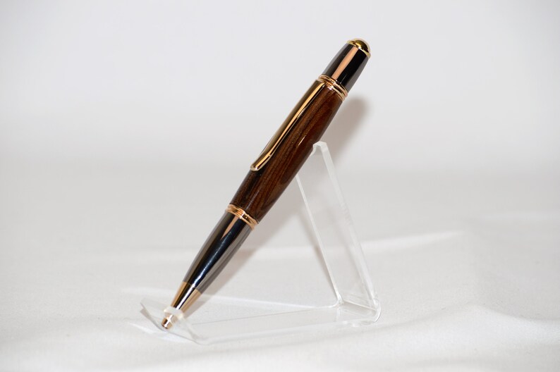 Puede incluir: Pluma de madera artesanal con cuerpo marr&oacute;n oscuro y detalles dorados. La pluma presenta un dise&ntilde;o elegante con punta negra y clip dorado. Se exhibe sobre un soporte de acr&iacute;lico transparente, mostrando su elegancia. Mide aproximadamente 14 cm.