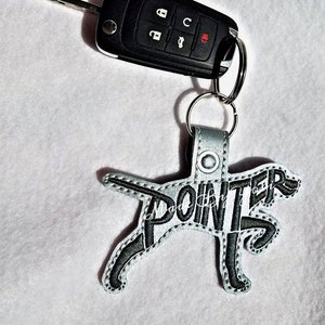 Op de afbeelding: Een zilvergrijze honden-sleutelhanger met het woord "POINTER" geborduurd in zwart. De sleutelhanger is bevestigd aan een zilveren sleutelring met een zwarte autosleutel.