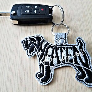 May include: Black and white dog silhouette keychain with the name "Waten" embroidered on it. The keychain is attached to a silver key ring.