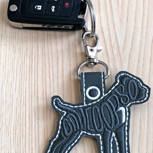 May include: A grey leather keychain featuring a black embroidered dog silhouette. The dog's outline is filled with the words "LOVE YOU" in a cursive font. The keychain is attached to a silver key ring and a black car key.