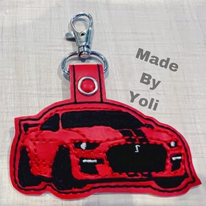 K&ouml;nnte beinhalten: Roter Leder-Schl&uuml;sselanh&auml;nger mit einem schwarzen gestickten Bild eines Ford Mustang. Der Schl&uuml;sselanh&auml;nger hat einen silbernen Metallschl&uuml;sselring und der Text "Made By Yoli" ist im Hintergrund sichtbar.
