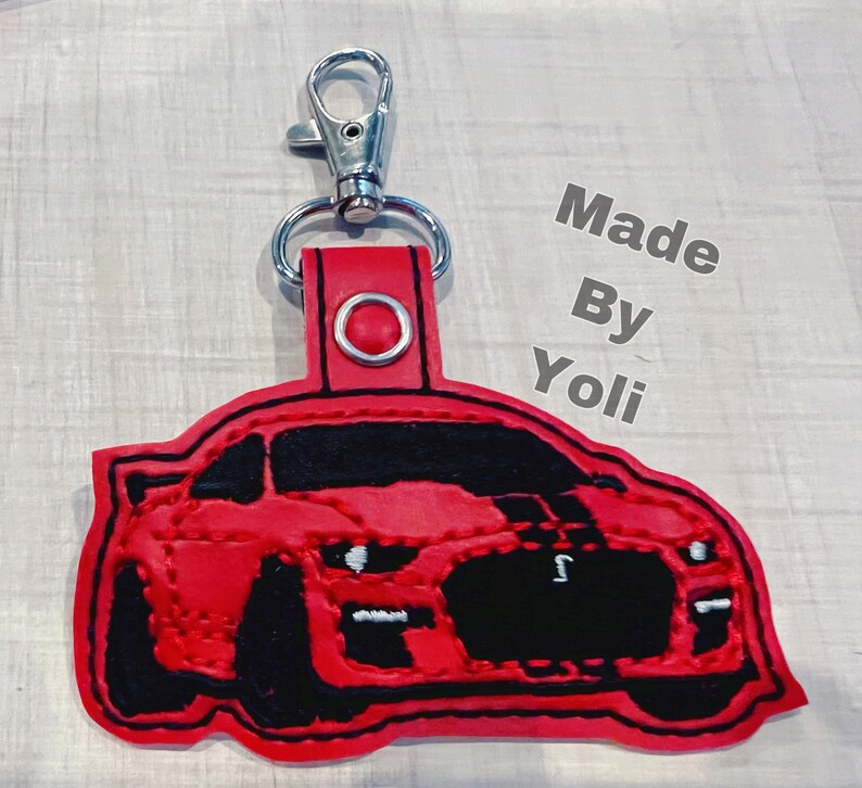 K&ouml;nnte beinhalten: Roter Leder-Schl&uuml;sselanh&auml;nger mit einem schwarzen gestickten Bild eines Ford Mustang. Der Schl&uuml;sselanh&auml;nger hat einen silbernen Metallschl&uuml;sselring und der Text "Made By Yoli" ist im Hintergrund sichtbar.