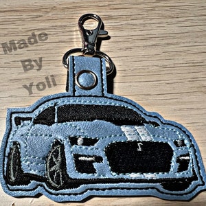 K&ouml;nnte beinhalten: Ein hellblauer Leder-Schl&uuml;sselanh&auml;nger mit einem schwarzen und wei&szlig;en gestickten Bild eines Ford Mustang. Der Schl&uuml;sselanh&auml;nger hat einen silbernen Metallverschluss.
