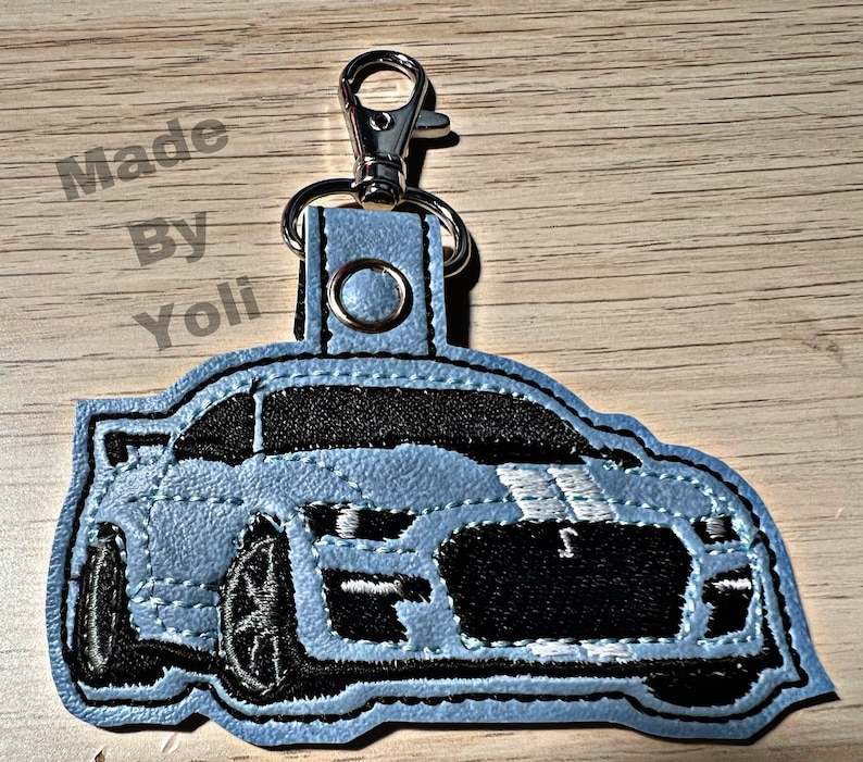 K&ouml;nnte beinhalten: Ein hellblauer Leder-Schl&uuml;sselanh&auml;nger mit einem schwarzen und wei&szlig;en gestickten Bild eines Ford Mustang. Der Schl&uuml;sselanh&auml;nger hat einen silbernen Metallverschluss.