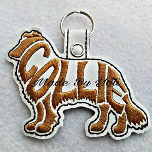 Könnte beinhalten: Weißer Leder-Schlüsselanhänger mit einer braunen gestickten Collie-Hundesilhouette. Die Silhouette des Hundes besteht aus dem Wort "Collie" in einer kursivschriftlichen Schriftart. Der Text "Made By Yoti" ist unter dem Hund gestickt.