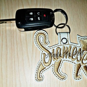 May include: White leather keychain with a gold embroidered cat silhouette and the word "Siamese" written inside the cat. The keychain is attached to a silver key ring.