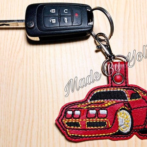 May include: Red embroidered car keychain with black details and a yellow stripe. The keychain is attached to a silver key ring and has the text "Made By Yoli" on the back.