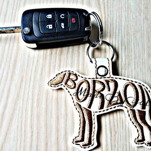 May include: A white leather keychain with a brown embroidered dog silhouette. The dog's silhouette is filled with the word "BORZOI" in brown embroidery. The keychain is attached to a silver key ring.