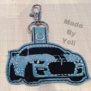 K&ouml;nnte beinhalten: Ein hellblauer Schl&uuml;sselanh&auml;nger aus Filz mit einem schwarzen gestickten Bild eines Autos. Der Schl&uuml;sselanh&auml;nger hat einen silbernen Metallring und einen Clip. Der Text "Made By Yoli" ist im Hintergrund sichtbar.